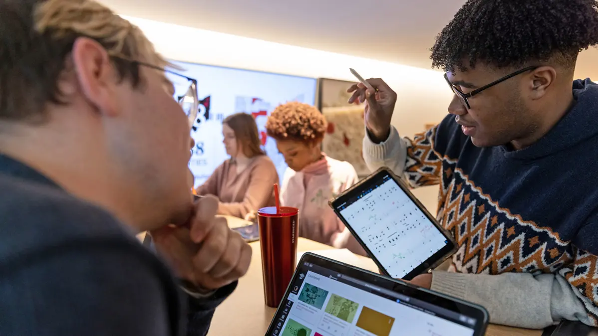 A young black man wearing glasses and a patterned sweater shows a fellow student a tablet computer with sheet music that he's made notations on. They're both engrossed by it.