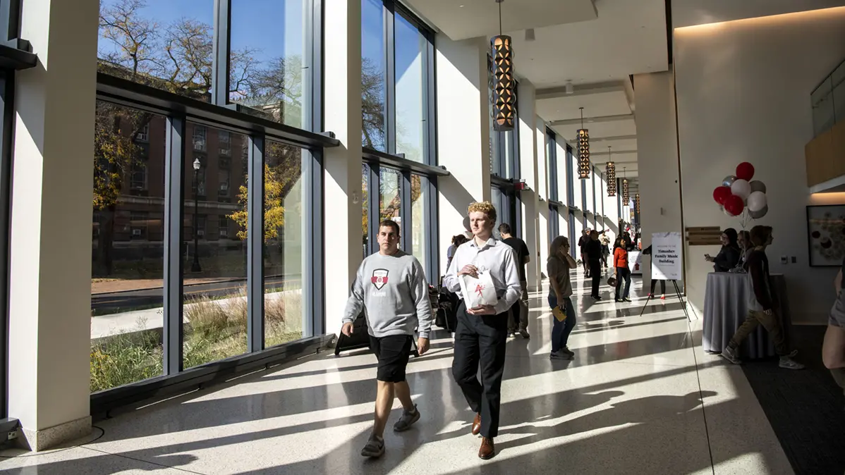 Students walk down a hallway where the late-afternoon sun streaming in through the wall of windows casts shadows on the floor.