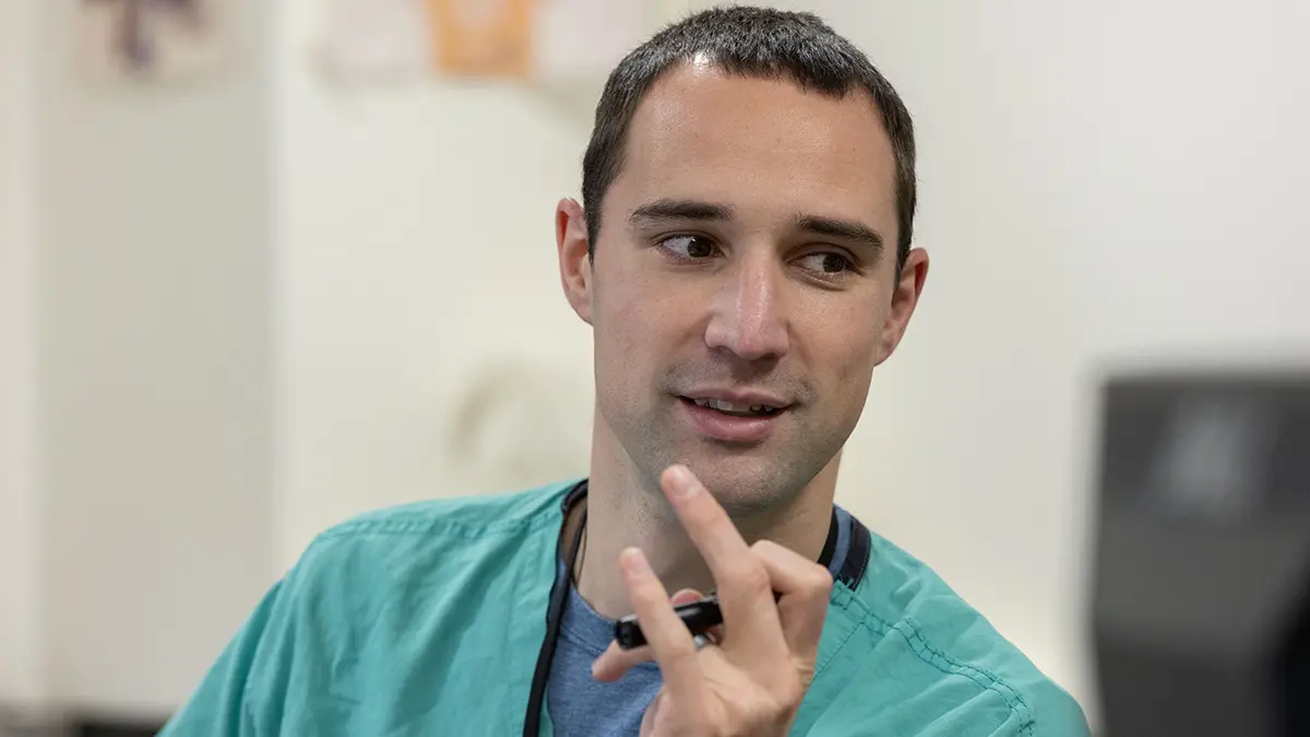 A white man with short dark hair and dark eyes smiles and twirls a pen as he looks at someone off camera. He’s wearing green scrubs and a lanyard.