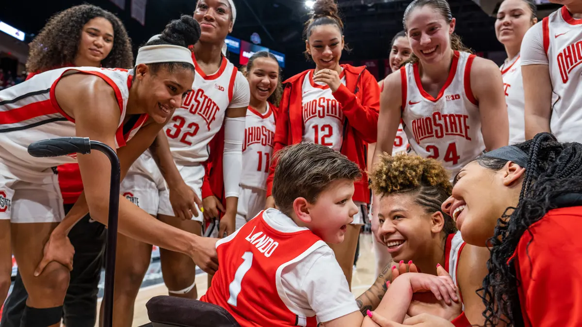 Ohio State women’s basketball players circle around a boy in a wheelchair, who is wearing a team and seen from behind. Most of the women are smiling or laughing.