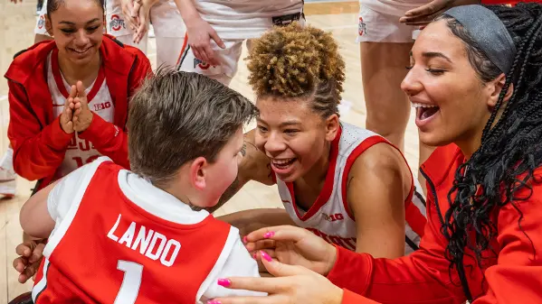 Ohio State women’s basketball players circle around a boy in a wheelchair, who is wearing a team and seen from behind. The women are smiling.