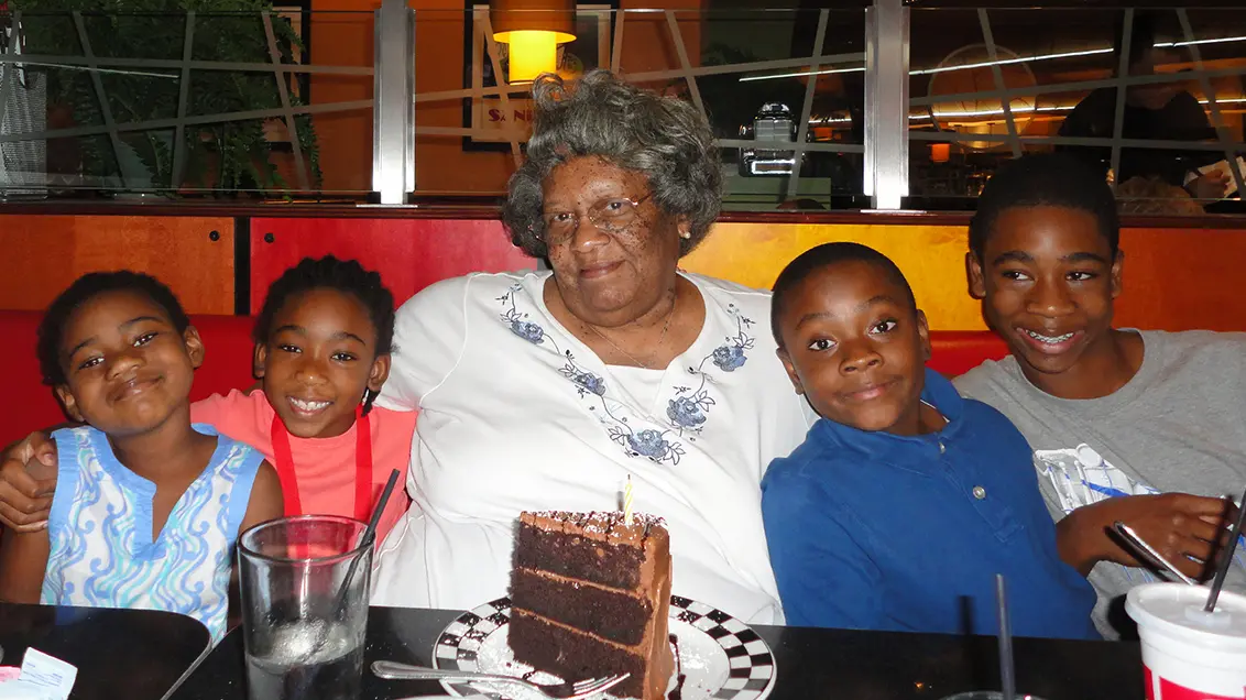 In a restaurant, a huge slice of chocolate cake sits on the table in front of a Black woman with gray hair. She is smiling proudly — looking a bit mischievous, too — has her arms around four preteen great-grandkids in the restaurant booth with her. The four are all smiling.