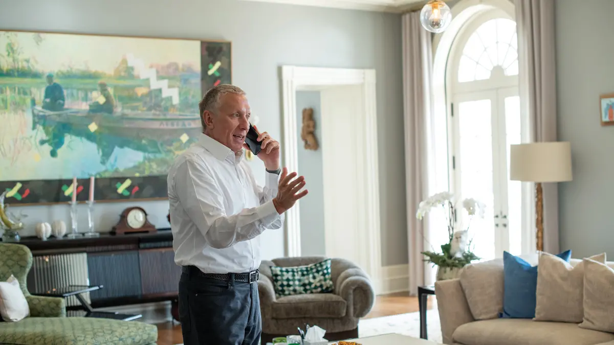 A man gestures while talking on his cellphone in his well-decorated living room. The upholstered couch and chairs seem cushy and reflect the colors from a large painting on the wall, a mix of impressionism and modern art.