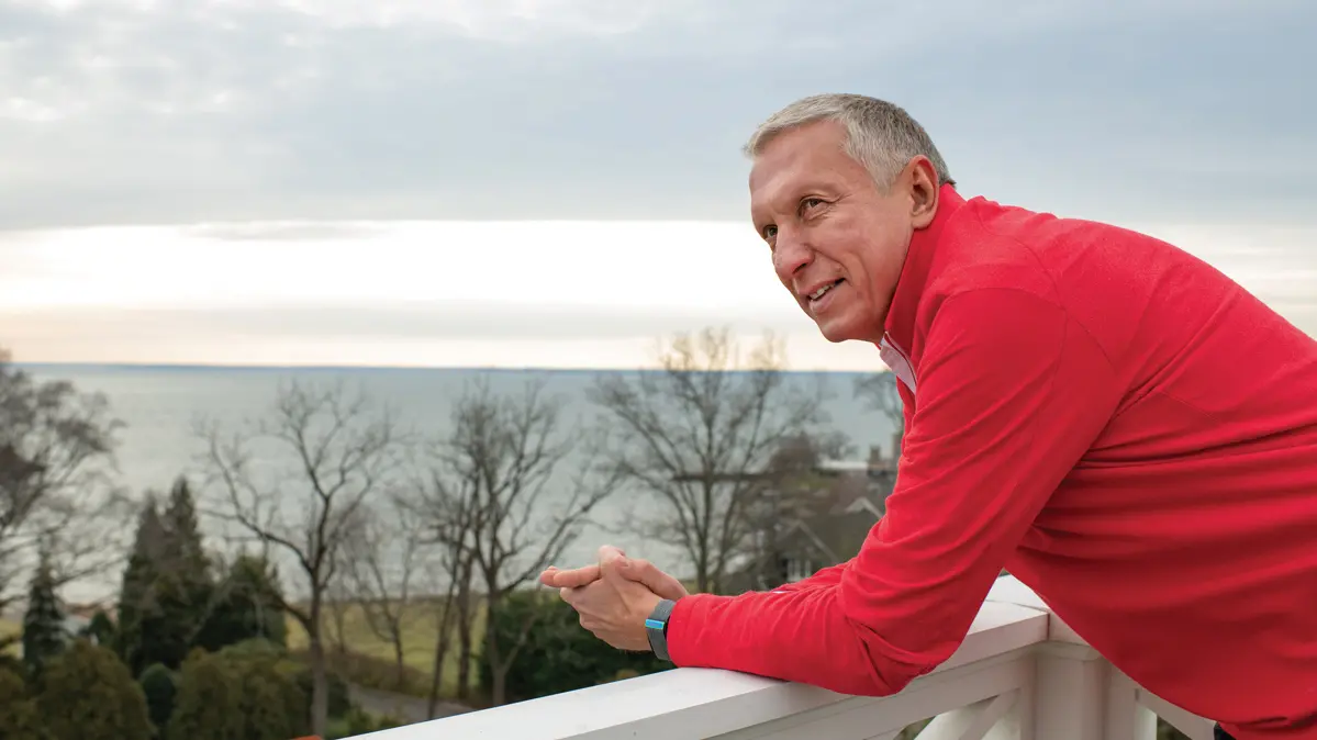 A white-haired man wearing a scarlet shirt leans on a railing on a roof-top porch with a view of the ocean. He has an interested, happy expression, as if he is looking, not at the pretty scene, but at someone talking next to him.