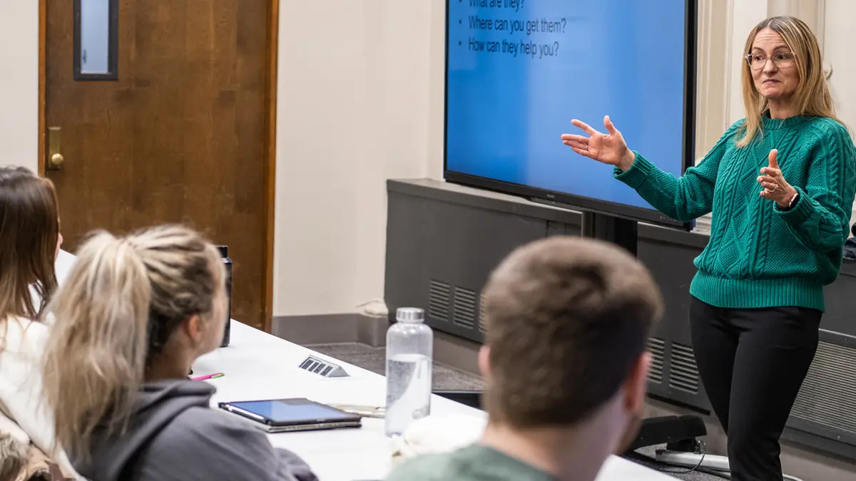 In an Ohio State College of Optometry class, students sit at a long table at the front of a classroom watching a teacher define mentoring. The teacher, a blond white woman wearing glasses, has her hands spread wider than her body, as if encompassing all of the students in the room. A screen behind her says “Coaching, Consultation & Mentoring—what are they?”