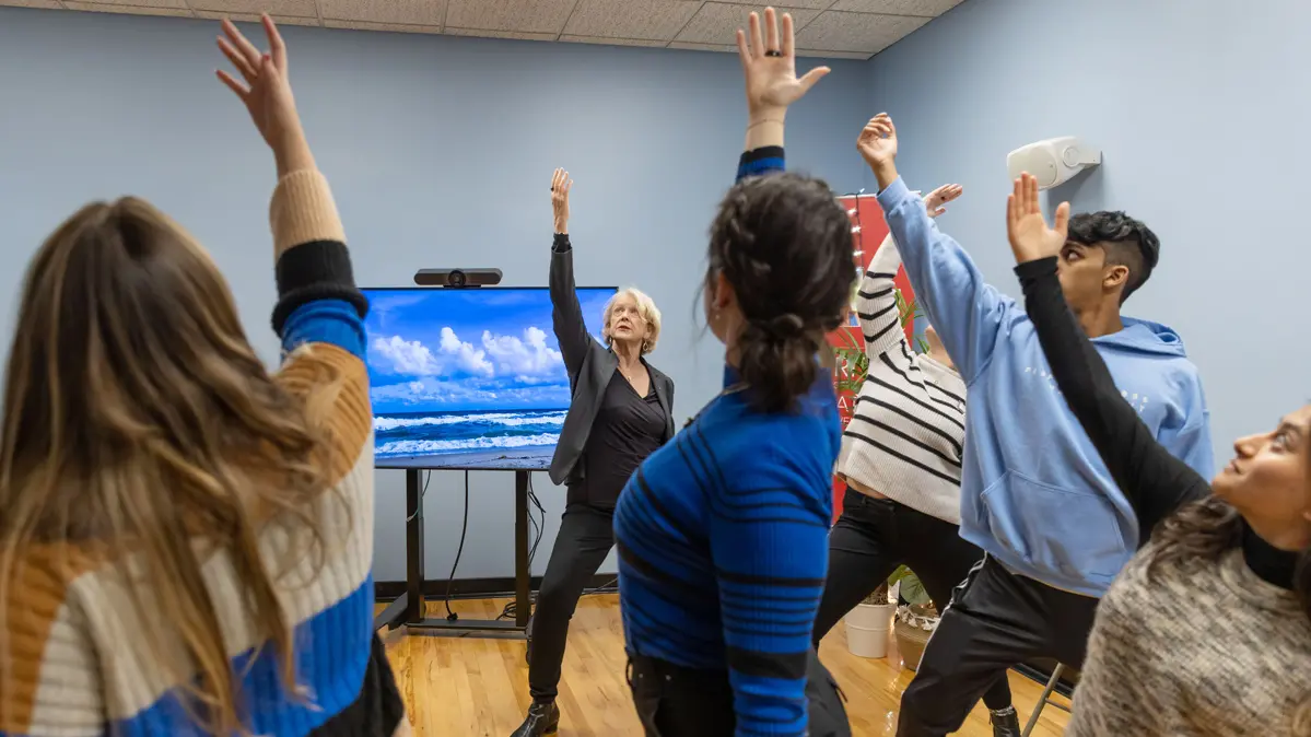 Five people stand in a loose circle, emulating the yoga pose of Professor Maryanna Klatt, an older white woman focusing on her stretch. They’re each leaning into their front leg and reaching up with the arm on that same side of their body. In the background is a large screen showing a pretty scene of waves rolling in the ocean and fluffy clouds.