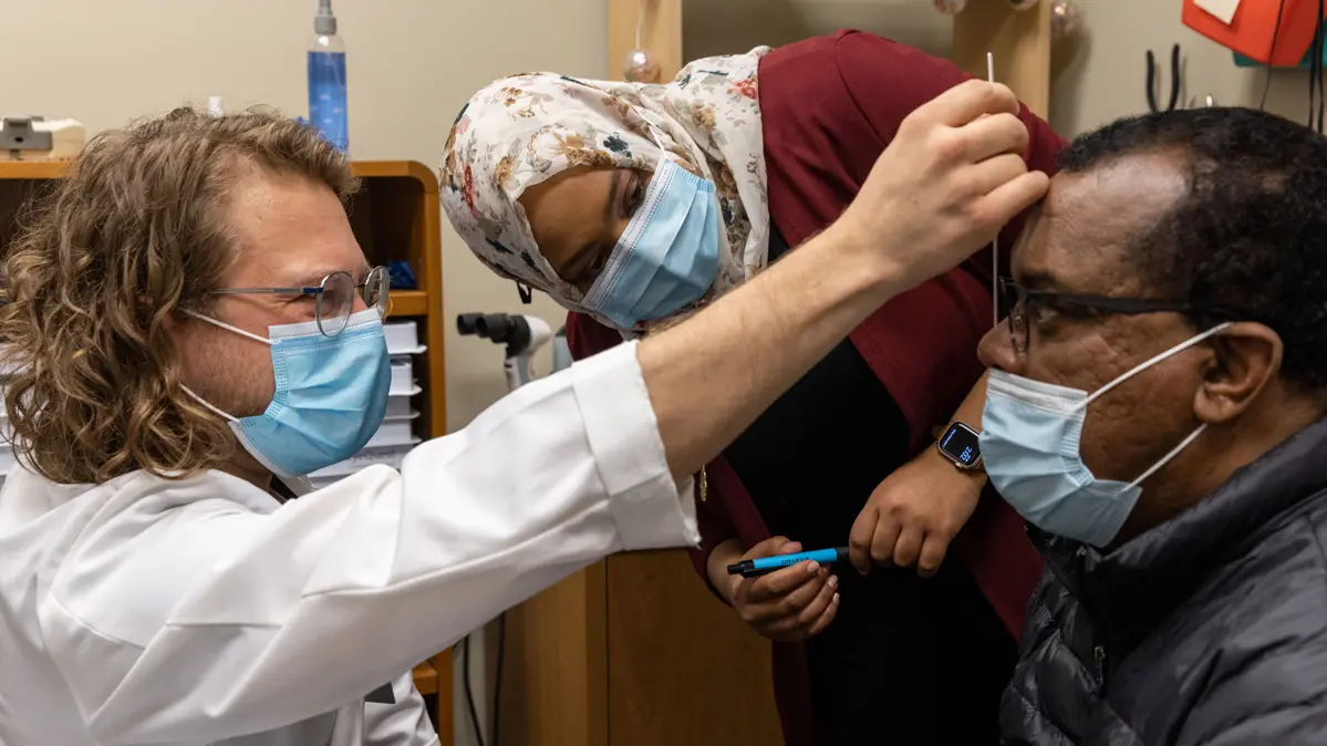 From left, a long-haired male student smiles and holds a measuring tool in front of a patient’s nose and forehead. His teacher, a Black doctor wearing a flowered headscarf, leans sideways to check on his technique and the patient, and the patient, a Black man wearing a winter coat and glasses, watches the measuring tool. All wear matching paper face masks.