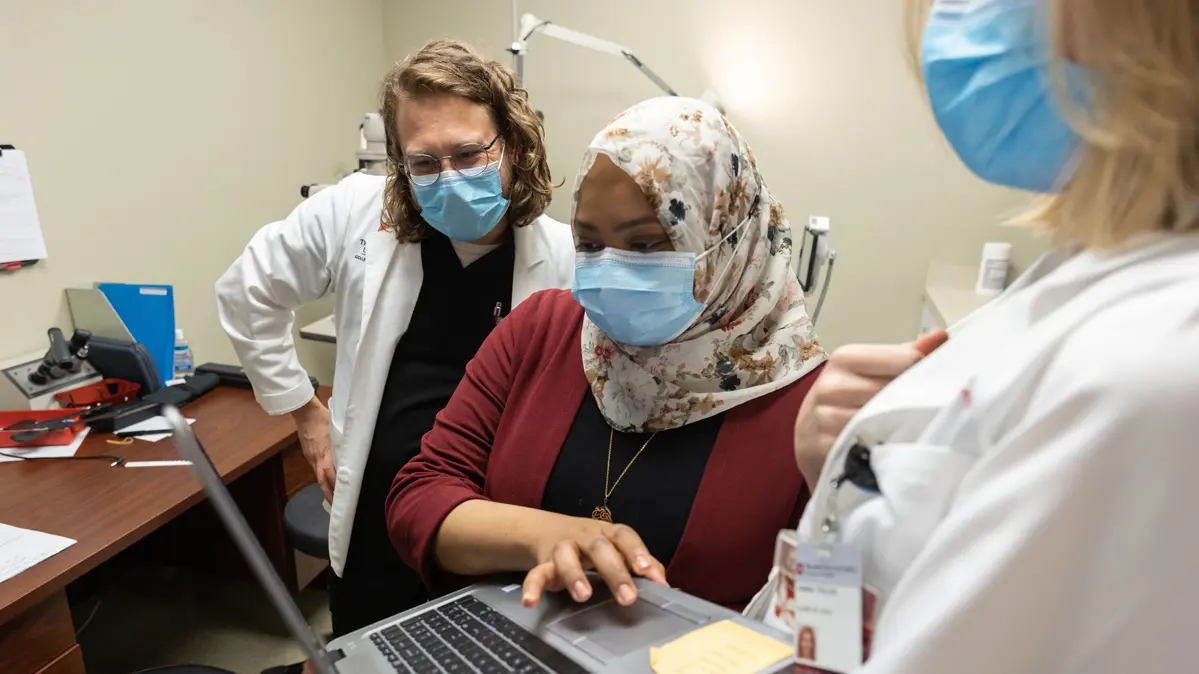 Standing in an optometry office, two students and their teacher, an optometrist, look intently at a laptop screen. Behind them are a desk and eye exam equipment.