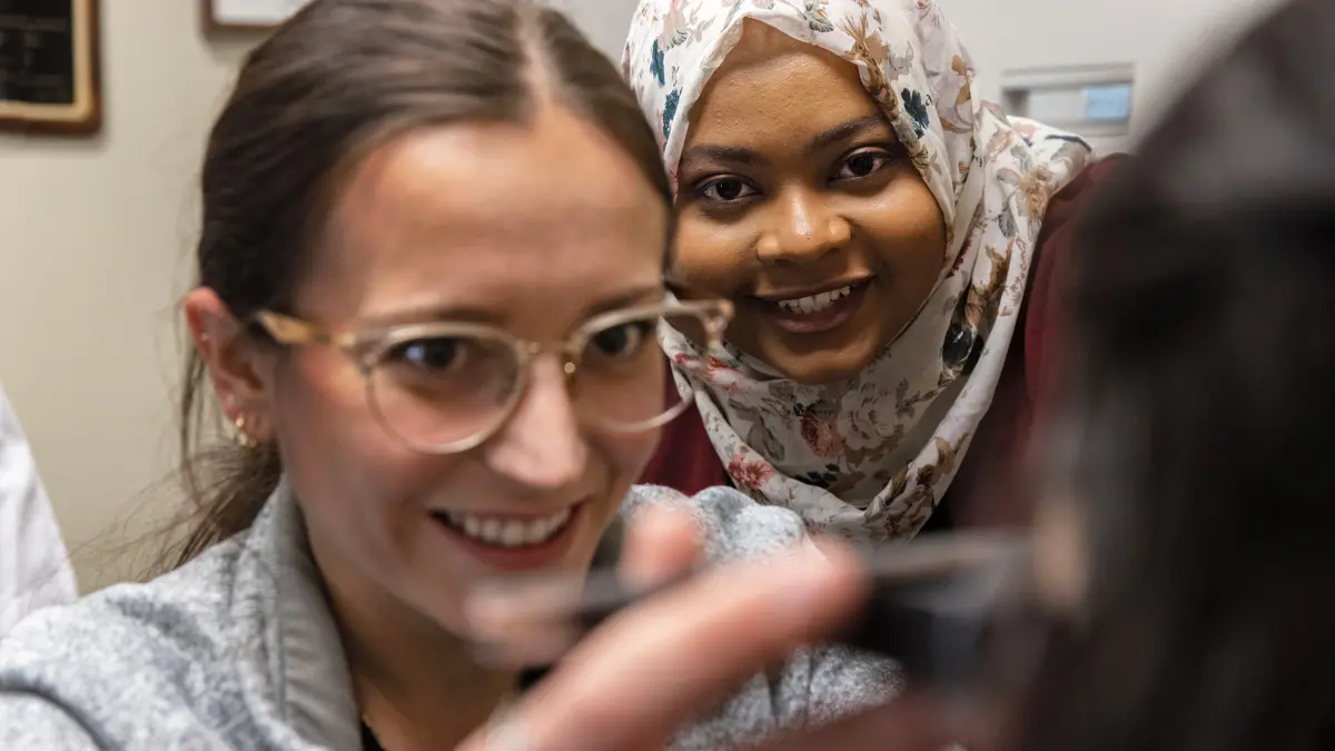 Two women chuckle as they walk through measuring another student’s eyes in an optometry office. The closer woman is a white college student wearing glasses and holding the instrument. She’s slightly out of focus. The woman in focus is her supervising doctor leaning in to check her work. The teacher is a Black woman wearing a flowered headscarf. 