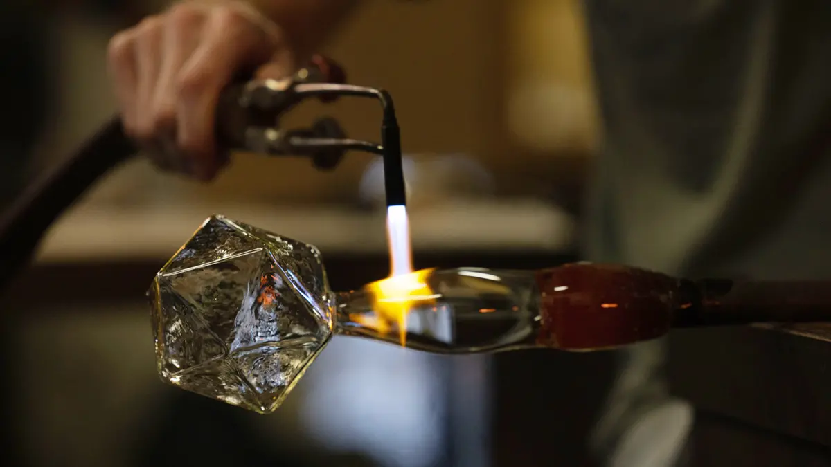 A blow torch is used on the glass stem of a many-sided clear glass piece, intended to represent a star for a celestial cluster. An artist holds the torch while stabilizing the glass work he just created. It is still attached to the pipe he used to blow air into the bubble of glass, before shaping.