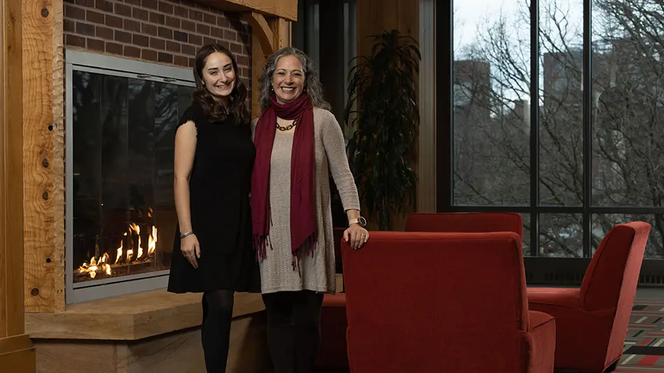 A young white woman poses with her mom in front of the fireplace at Longaberger Alumni House. They both wear dresses and big smiles, and have similarly wavy hair and stances.