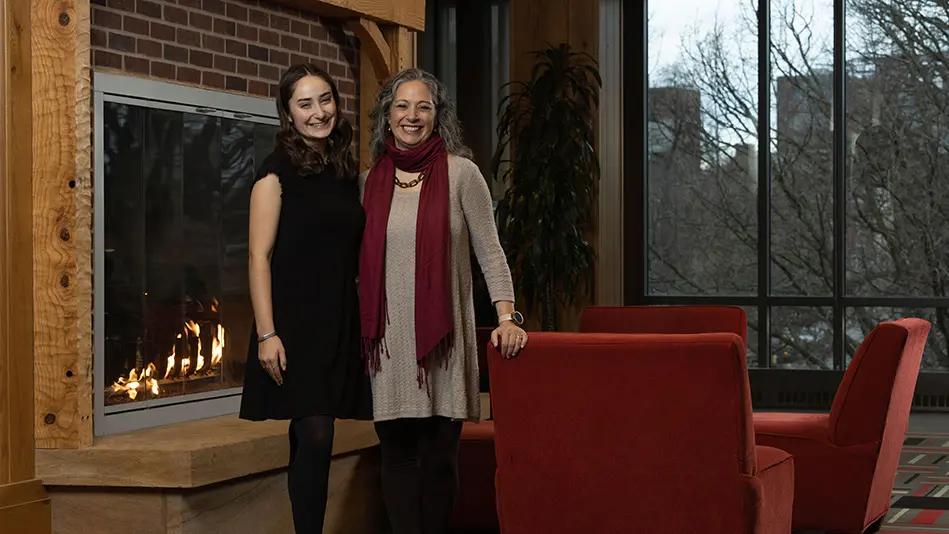 A young white woman poses with her mom in front of the fireplace at Longaberger Alumni House. They both wear dresses and big smiles, and have similarly wavy hair and stances.