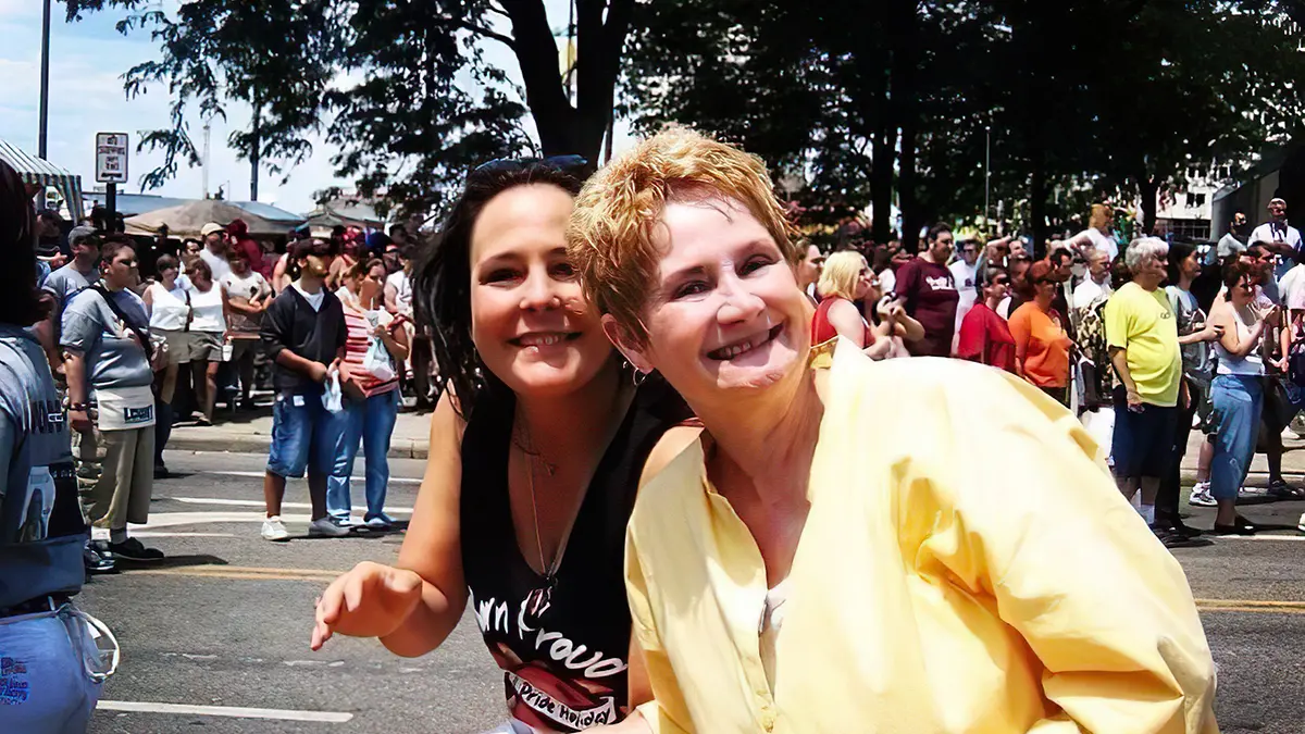 Two white women, one with short blond hair and the other with long dark hair, lean into the photo as parade-watchers line the street behind them. The women’s smiles say they are happy and proud.