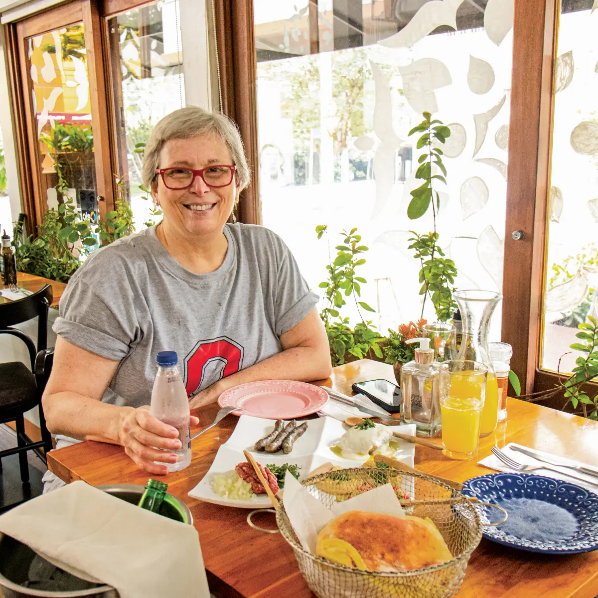 A woman smiles at a restaurant table. Her scarlet glasses match the color of the Block O on her T-shirt. Sunlight streams in windows behind her, plants grow on the sill, and colorful plates sit on her table awaiting the food.