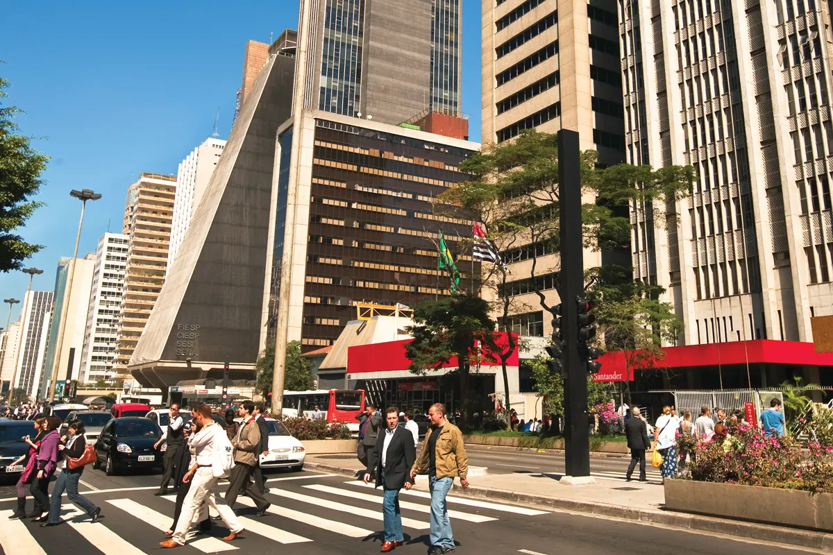 Skyscrapers line a street that has flower planters dividing the two directions of traffic. Pedestrians are shown crossing at a crosswalk. Bright blue sky peaks from behind the buildings.