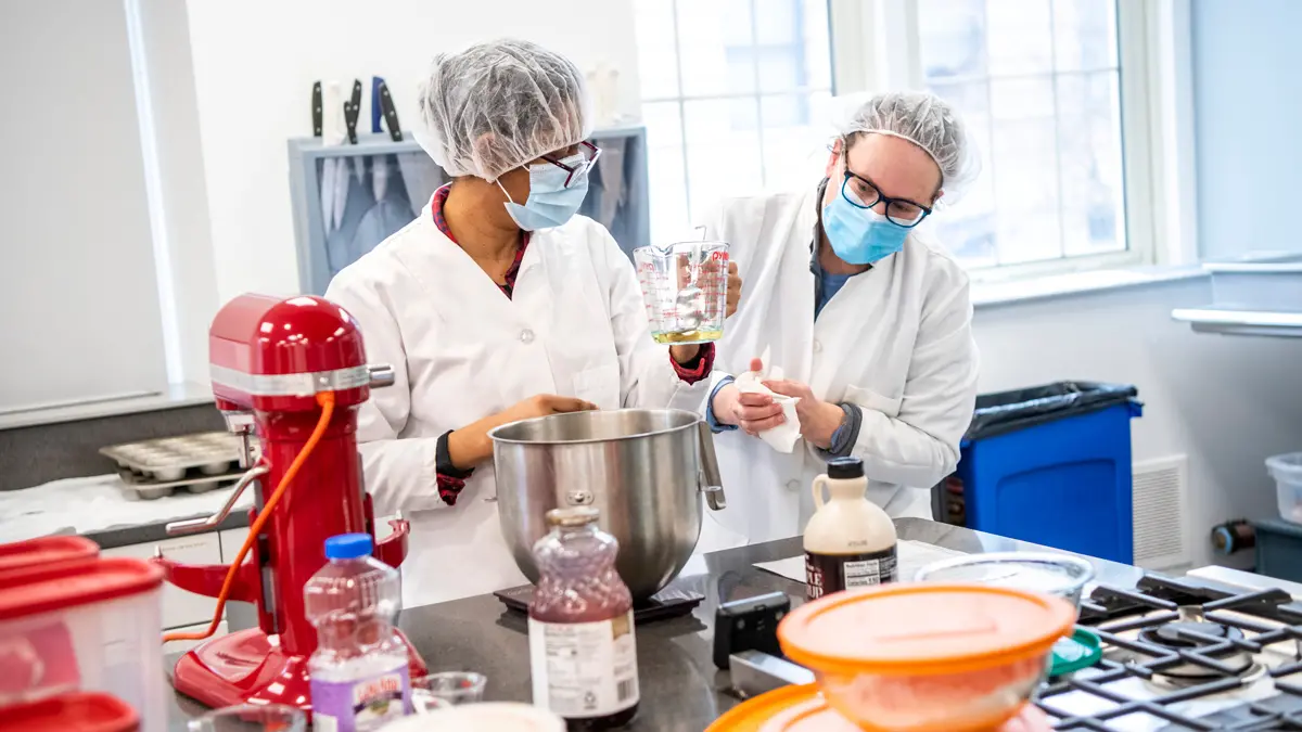 In a light-filled industrial kitchen, Two women wearing paper hair covers, face masks and glasses measure liquid to add to a mixing bowl. Also on their work table are a KitchenAid mixer, bottles of juice, oil and maple syrup and various other filled and capped bowls of ingredients