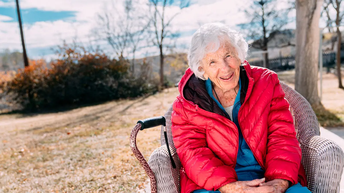 A senior woman with white hair smiles broadly and sits outdoors in a wicker chair wearing a red puffy winter coat. Her cane is by her side.