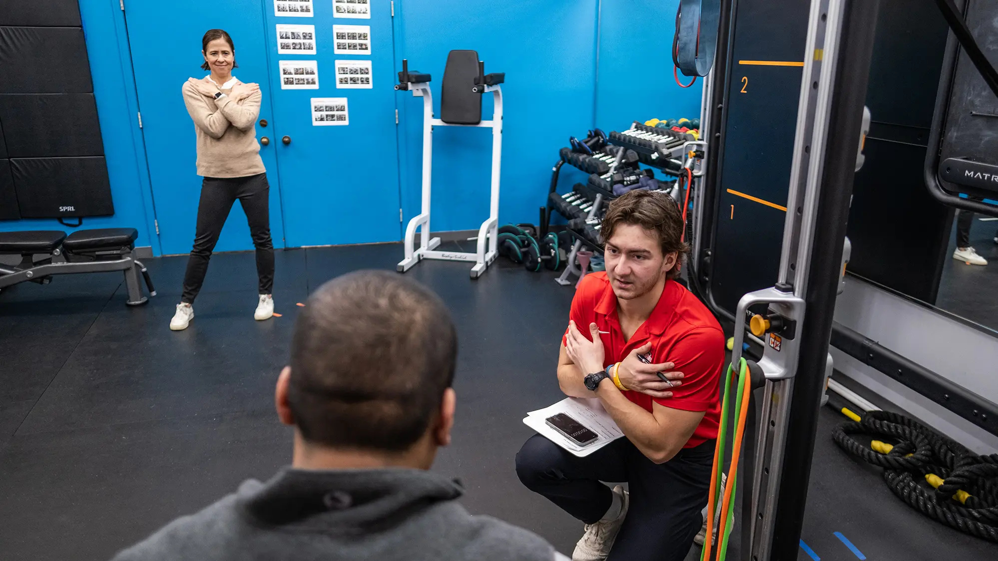 In a gym, a man sits with his back to the camera. Facing him (and by default, us) are a young man on the right side of the photo who is crouching, talking and crossing his arms over his chest, and further back and to the left, a smiling woman standing and making the same gesture.