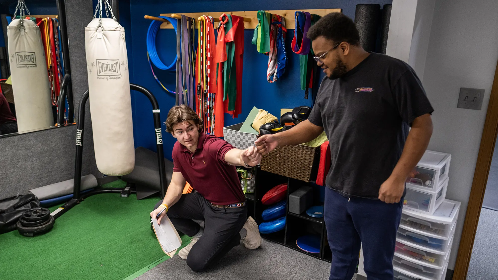 At a gym where a punching bag and pegs with exercise bands hang along a wall behind them, a young man crouching reaches up to fist bump a young man in sweats reaching down to make contact.