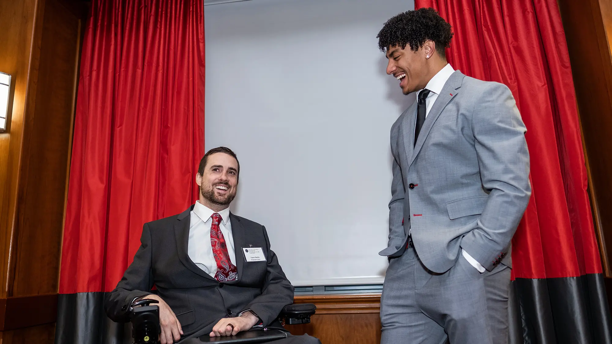 A white man seated in a wheelchair talks with a black man standing next to him and looking at him. Both laugh as they chat in their distinguished-looking suits, which say they’re at an important event.