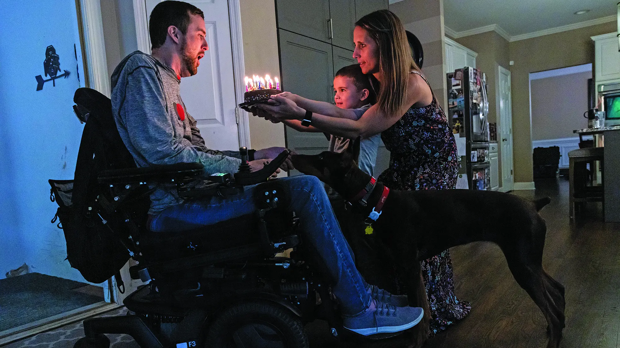 Megan, a white woman with long hair and wearing a long dress, crouches as she helps her 5-year-old son hold a cake ablaze with candles out to her wheelchair-bound husband, Tyson Gentry. He’s taking a big breath in, getting ready to blow out the candles, and the family dog is leaning forward to be part of the group.
