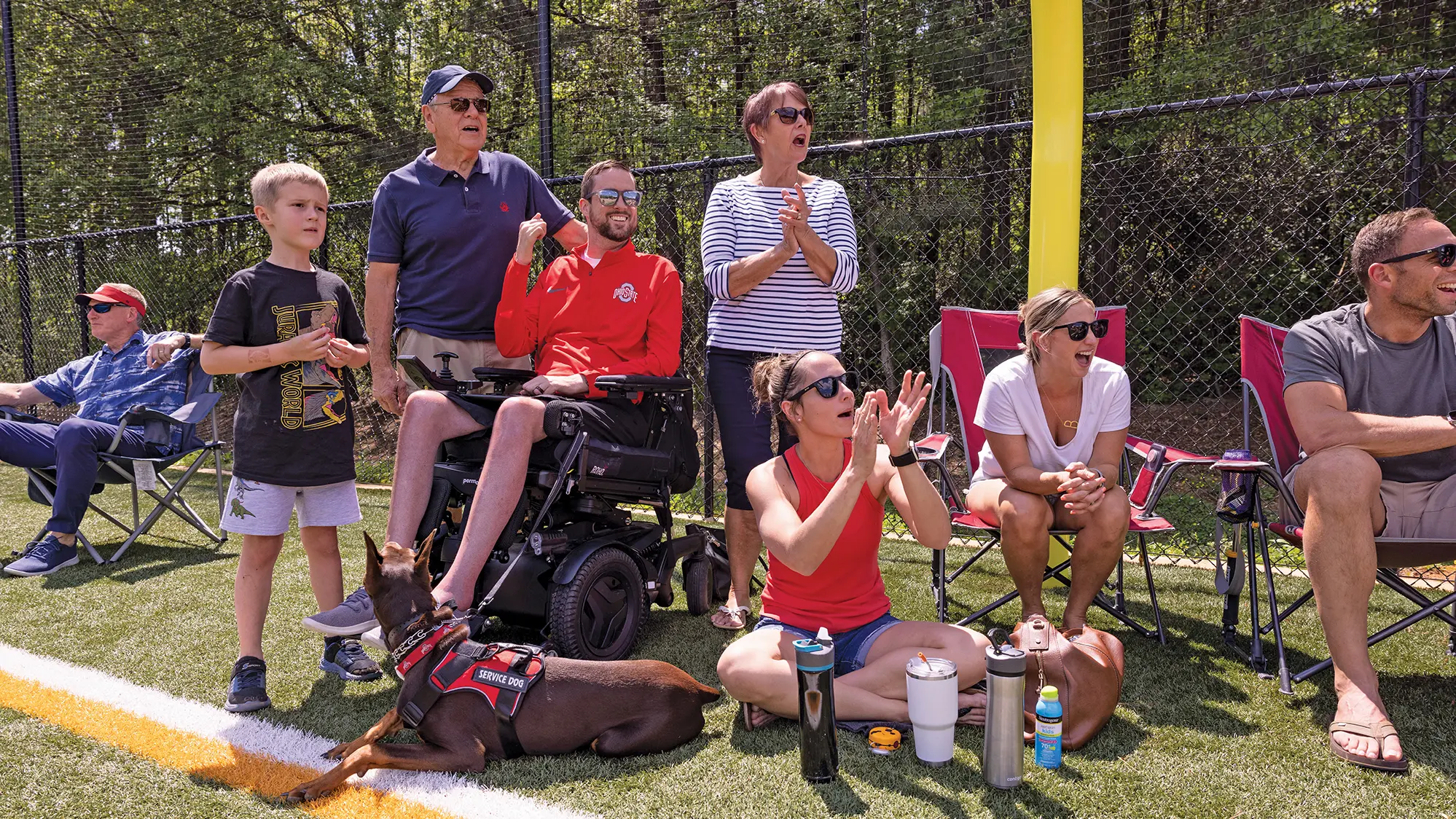 On a sunny day, spectators cheer from along a field sideline — some stand, some sit in chairs and Megan sits in the grass, beside the family Doberman, which wears a service dog vest. Everyone is cheering.