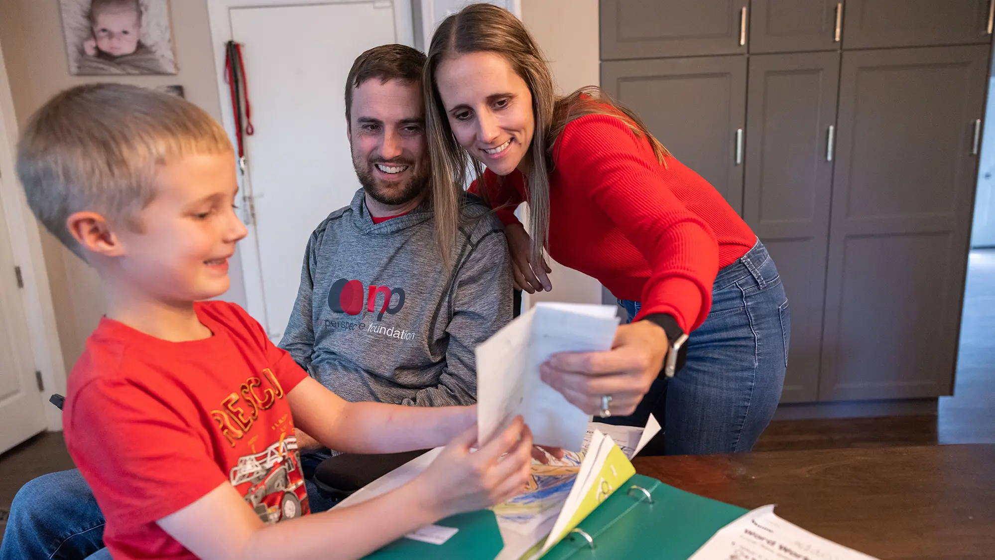 A young boy stands at a table holding up a drawing and showing it to his parents. His dad is in a wheelchair and his mom leans close to his dad, so they can look at the artwork together. The picture is facing away from the camera.