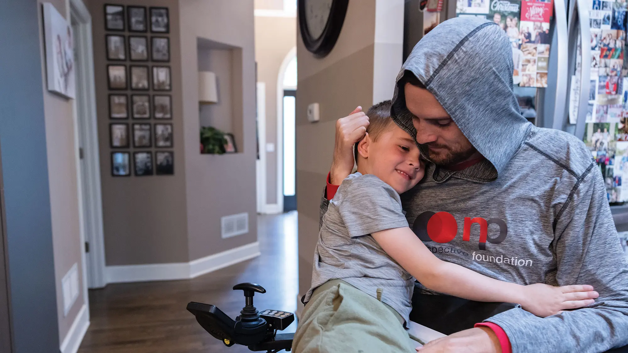A young boy smiles as he sits on the lap of his dad, Tyson Gentry, and hugs him. Tyson, a white, bearded, lanky man, has the arms of his wheelchair spread wider than usual and has the hood of his sweatshirt up. They’re in their home, where the walls and fridge in the background are covered in family photos.