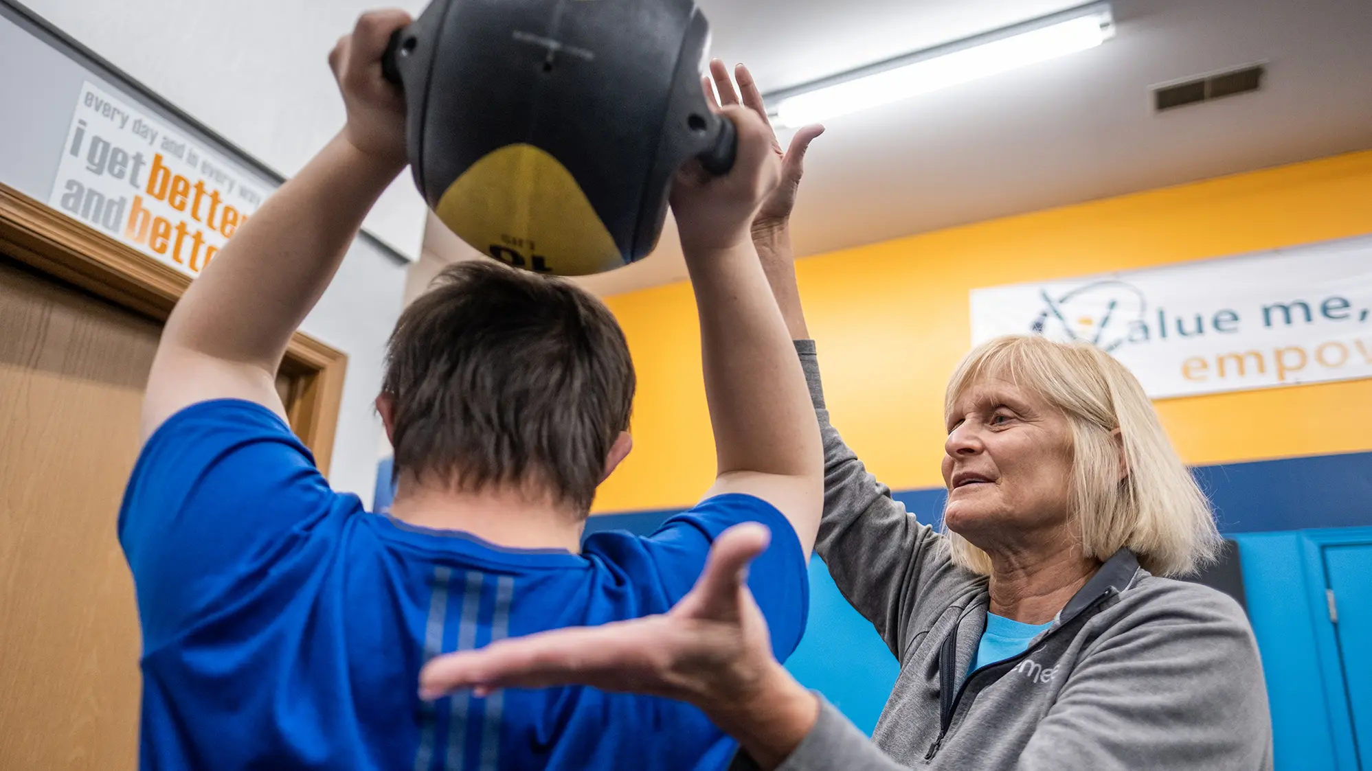 An older woman with blond hair vaguely smiles as she stands at the side of a man with intellectual or developmental disabilities lifting a medicine ball above his head. She's spotting him and as such, has her hands up and ready to grab the ball if he loses control. He can only be seen from behind.