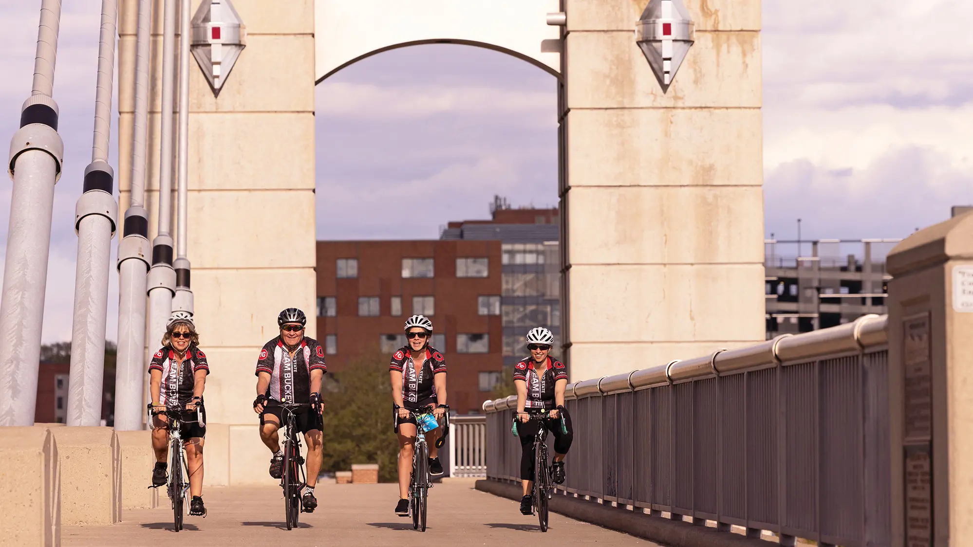 Biking across the Lane Avenue bridge, on the west side of campus, a family of four wears bike helmets, Team Buckeye jerseys, sunglasses and smiles. The four are a mom, dad and their two daughters.