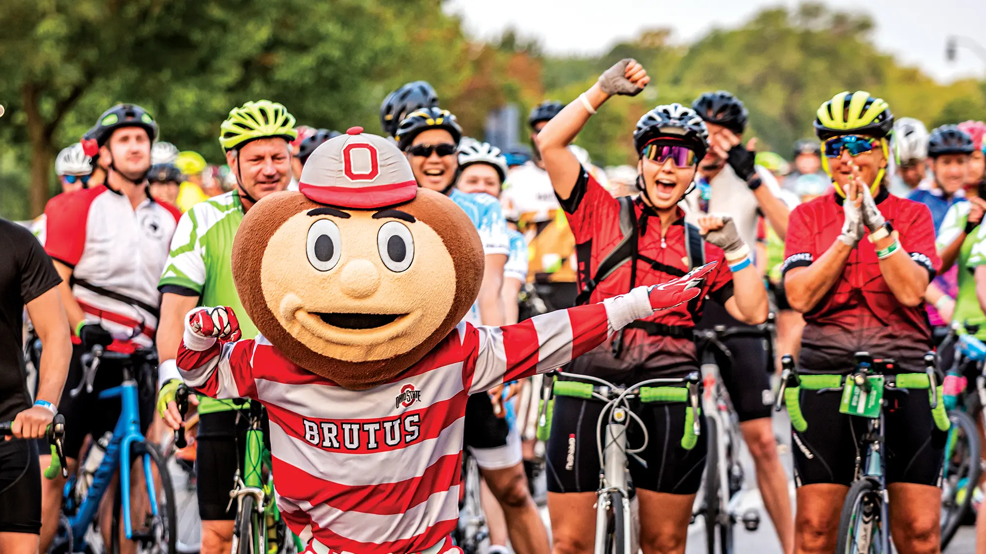Ohio State’s mascot Brutus Buckeye—who has big head designed to look like a buckeye nut and a regular-sized body wearing a striped, long-sleeved polo shirt—strikes a pose in front of a crowd of cheering riders, each standing over their bicycles and getting ready to start the Pelotonia ride.