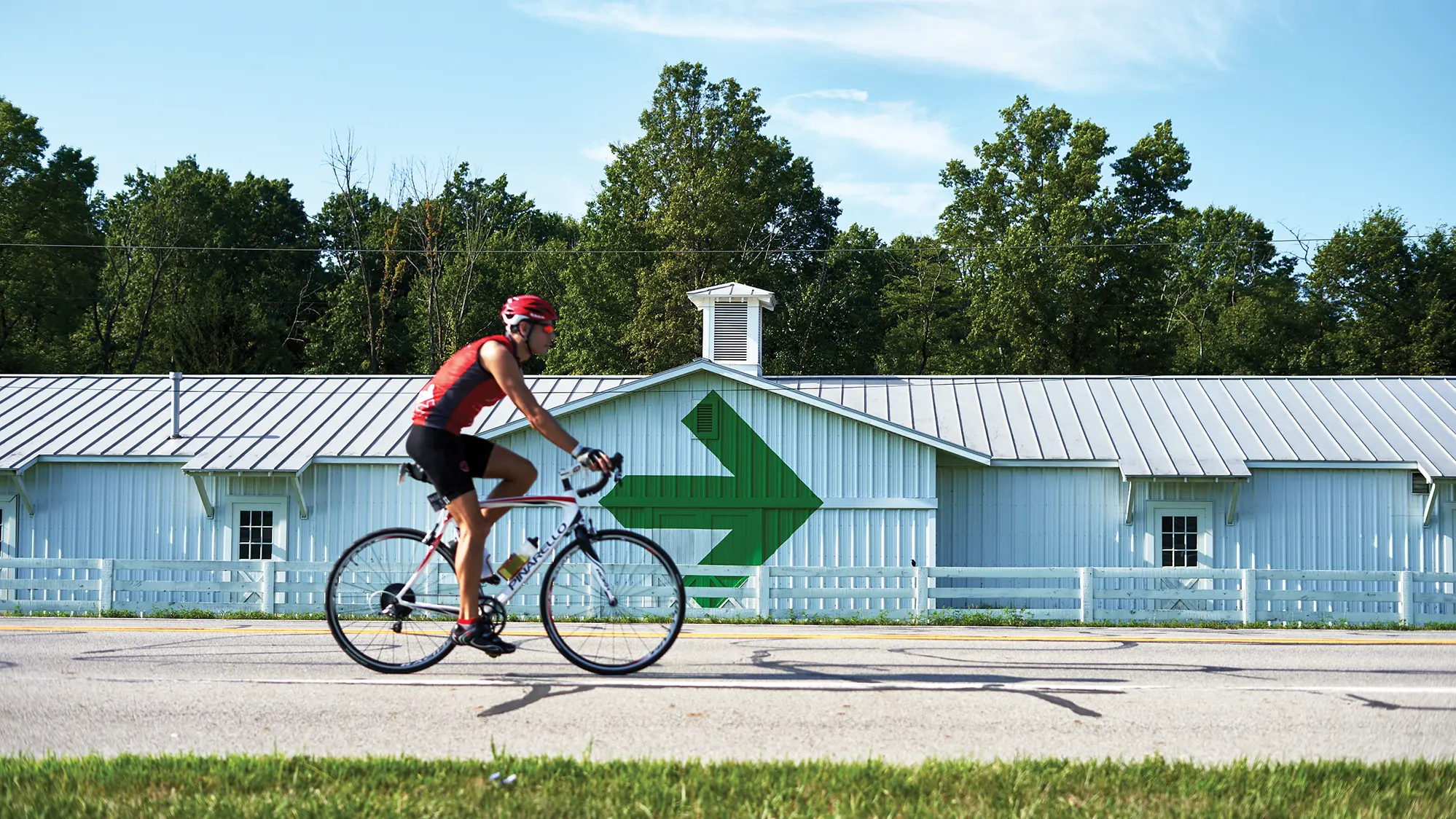 A lone male bicyclist rides past a well-cared for barn marked with a giant Pelotonia arrow, the ride’s logo. A short painted fence separates the road from the barn.