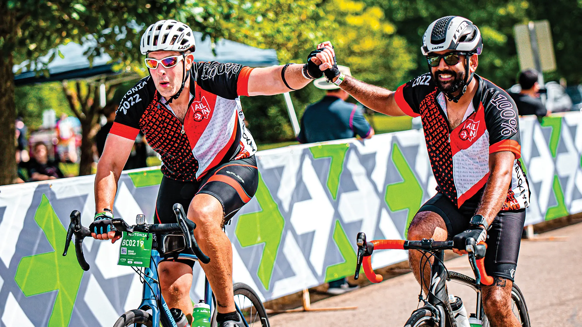 As they ride along a makeshift wall covered in Pelotonia arrow logos, two men on bikes high five. They’re likely near the finish line, and they both wear helmets, sunglasses and bicycle gloves. The man on the left is white and has an expression that says he is concentrating. The bearded man on the right looks to be of Indian descent and is smiling.