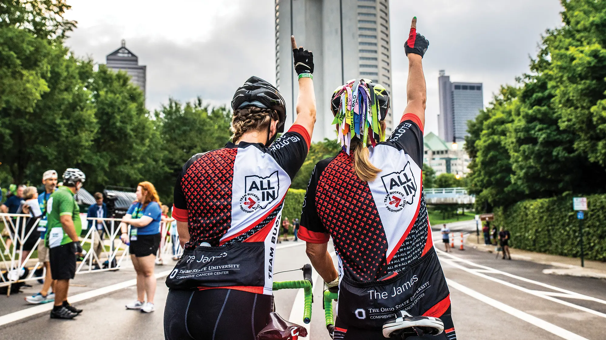 Two women raising their right hands high point to the sky, or make a number-one symbol. They’re seen from behind. The one on the left has intricately braided hair and a helmet, and the one on the right has a bike helmet trailing more than a dozen multicolored ribbons with writing on each. Far in front of them, downtown skyscrapers can be seen.