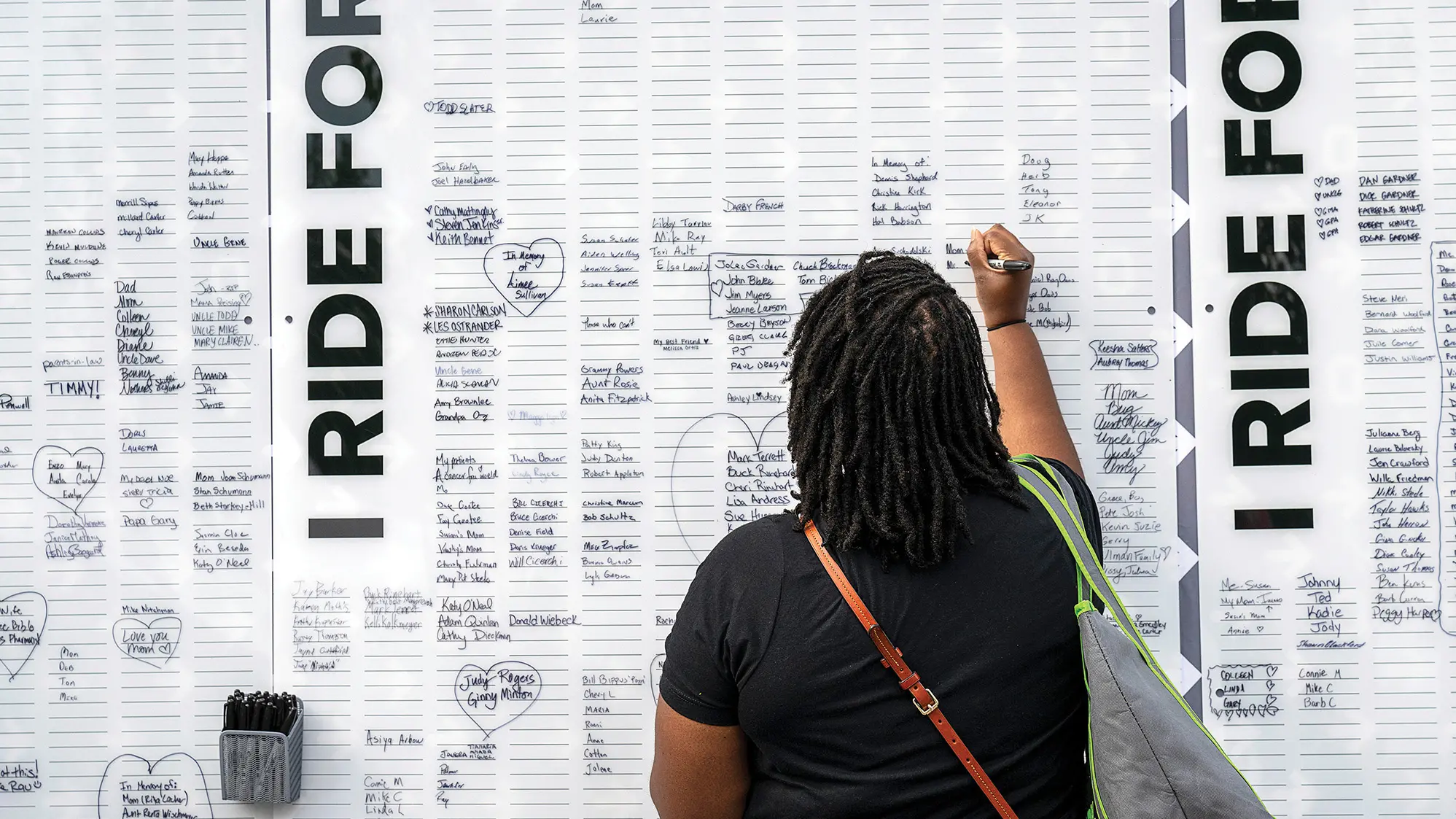 A black woman stands before a big white board posted upright, as if on a wall. It’s covered in short lines — many filled in with handwritten names — and on the side, it says, “I Ride For.” The woman has a marker  and is adding a name to the board.