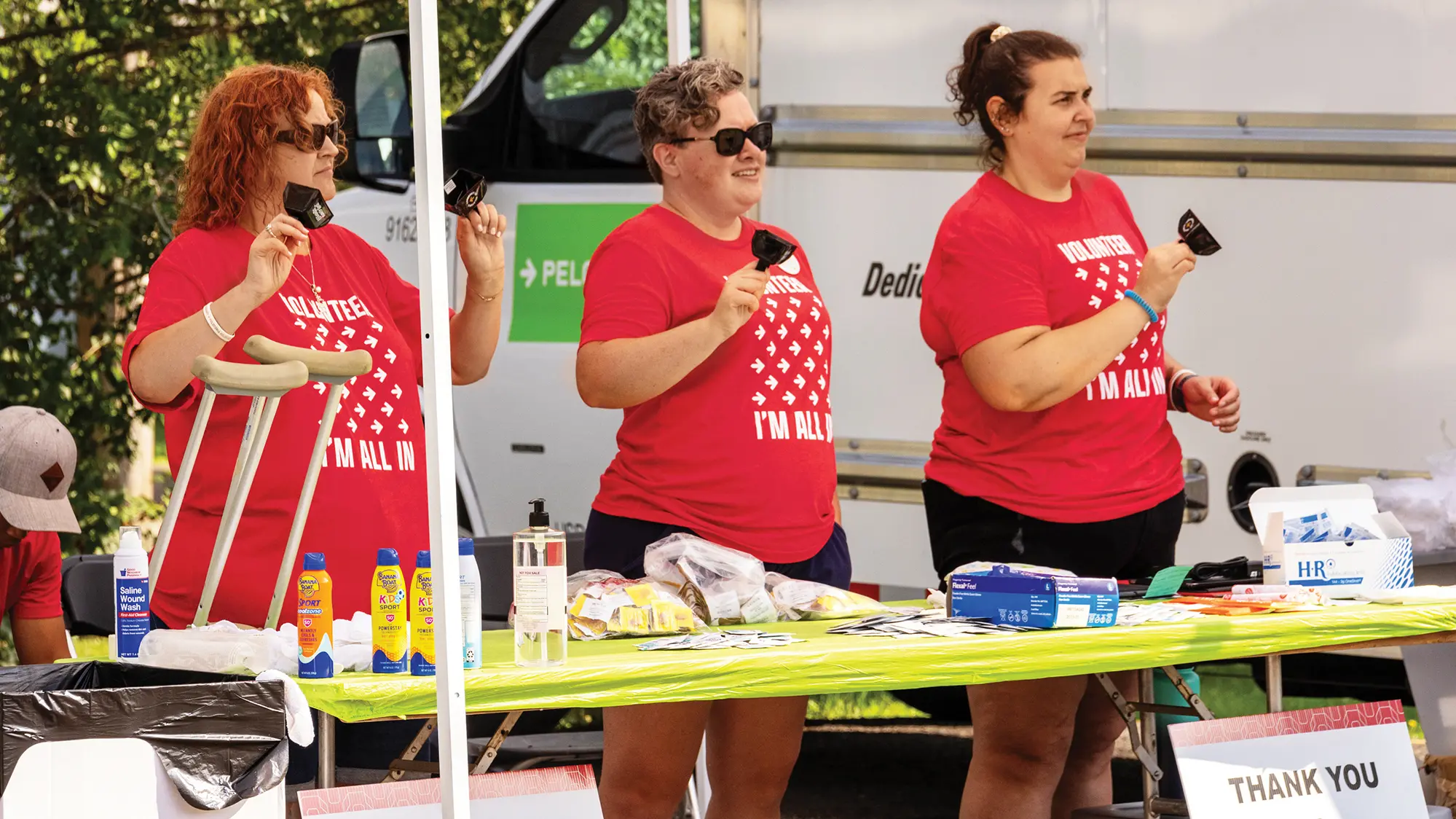 Three smiling white women stand behind a table stocked with first aid gear and sunblock lotion. Each woman rings a bell with her right hand, held about shoulder height, and the first woman has a second bell in her other hand. The bells are short and squat.