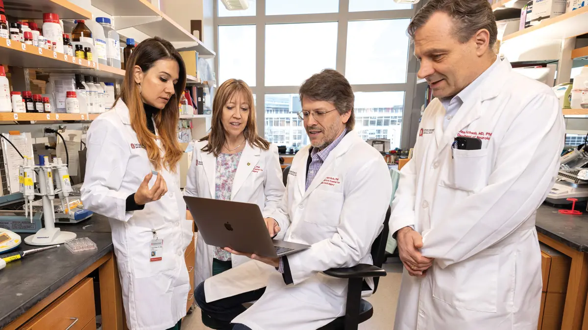 Four researchers wearing lab coats listen and talk in a room filled with shelves of medications. Two are men and two are women.
