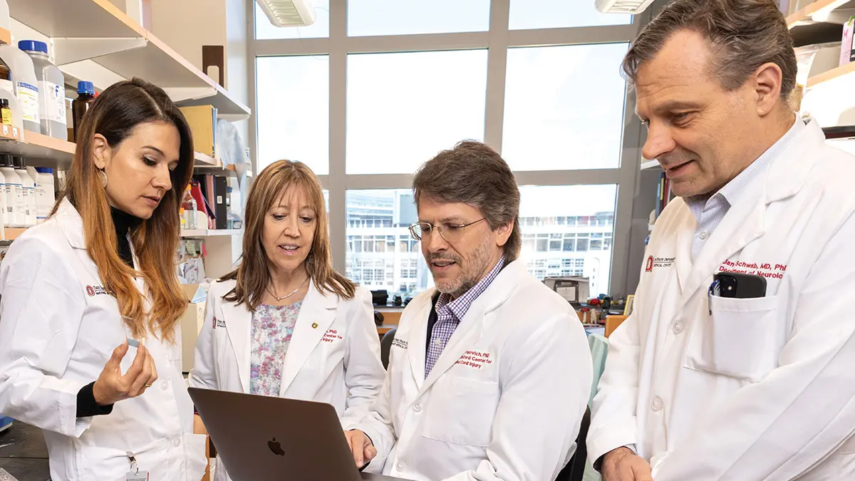 Four researchers wearing lab coats talk in a room filled with shelves of medications. Two are men and two are women.