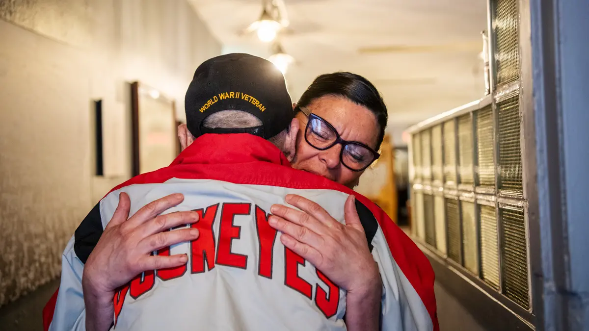 Two people hug. The man’s back is to the camera; his jacket says “Buckeyes” across his shoulders. The woman hugging him has both hands on that word and her eyes are closed as she gives him a squeeze.