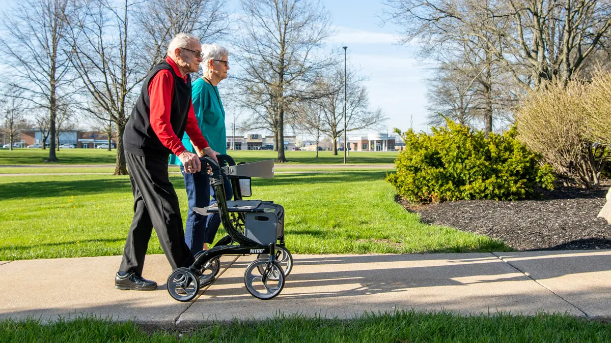 Passing a pretty portion of campus, while trees are just beginning to show their spring buds but the grass already has greened up, Lashey uses a rolling cart that provides extra support while he walks. (It looks like it could double as a seat but is not a traditional wheelchair.) His girlfriend walks beside him. They’re both wearing glasses and looking ahead.