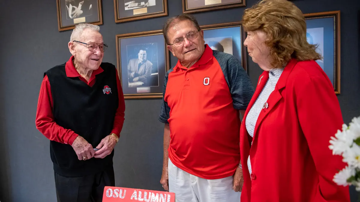 Behind a sign on a table that reads “OSU Alumni Club-Marion Co.” and a wrapped plate of treats, an older husband and wife in Ohio State gear chat with Lahey at an event. They’re making eye contact as they smile and listen to one another.