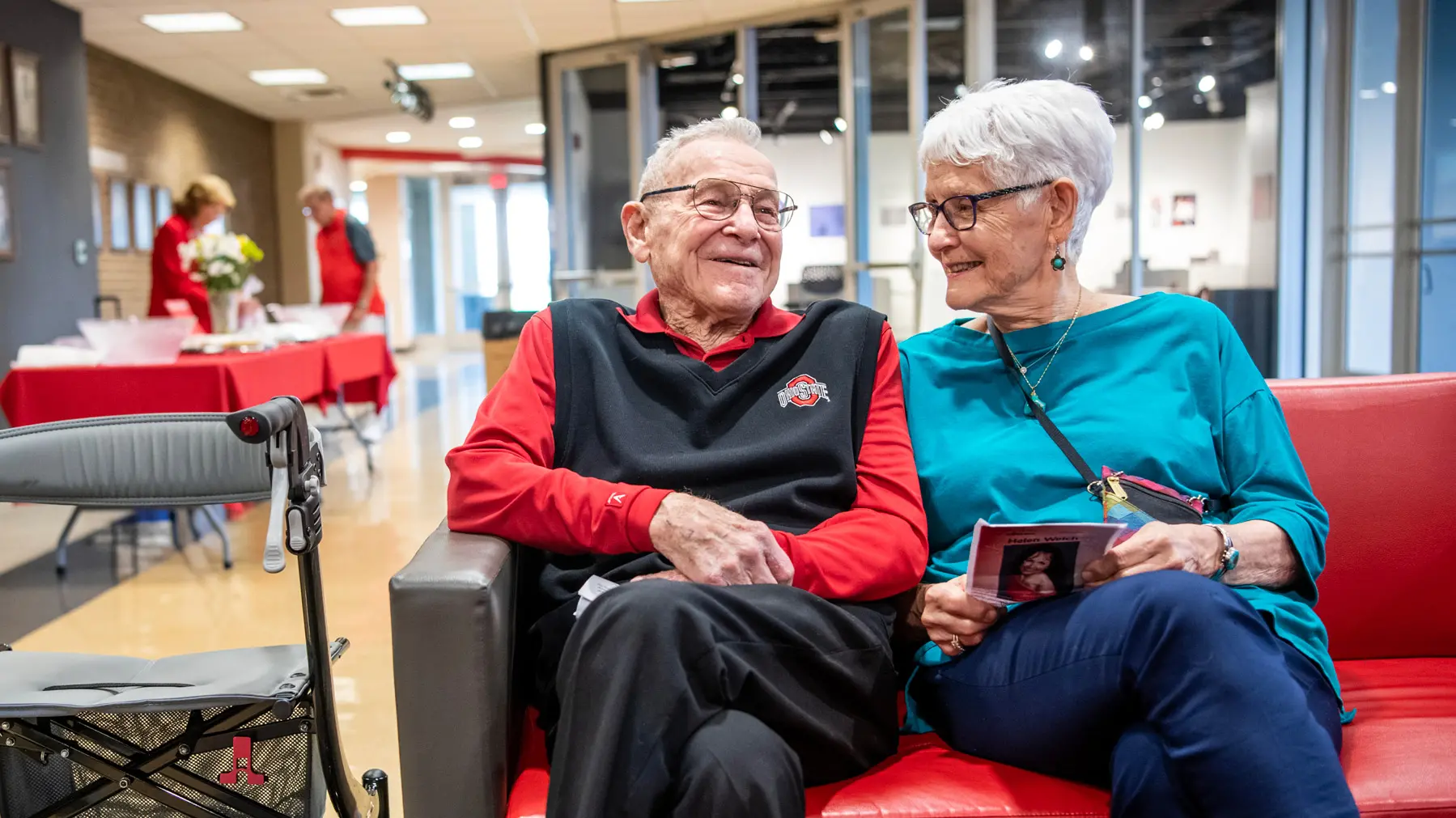 A white-haired couple sit together on a couch, smiling as they chat and lean against each other. 