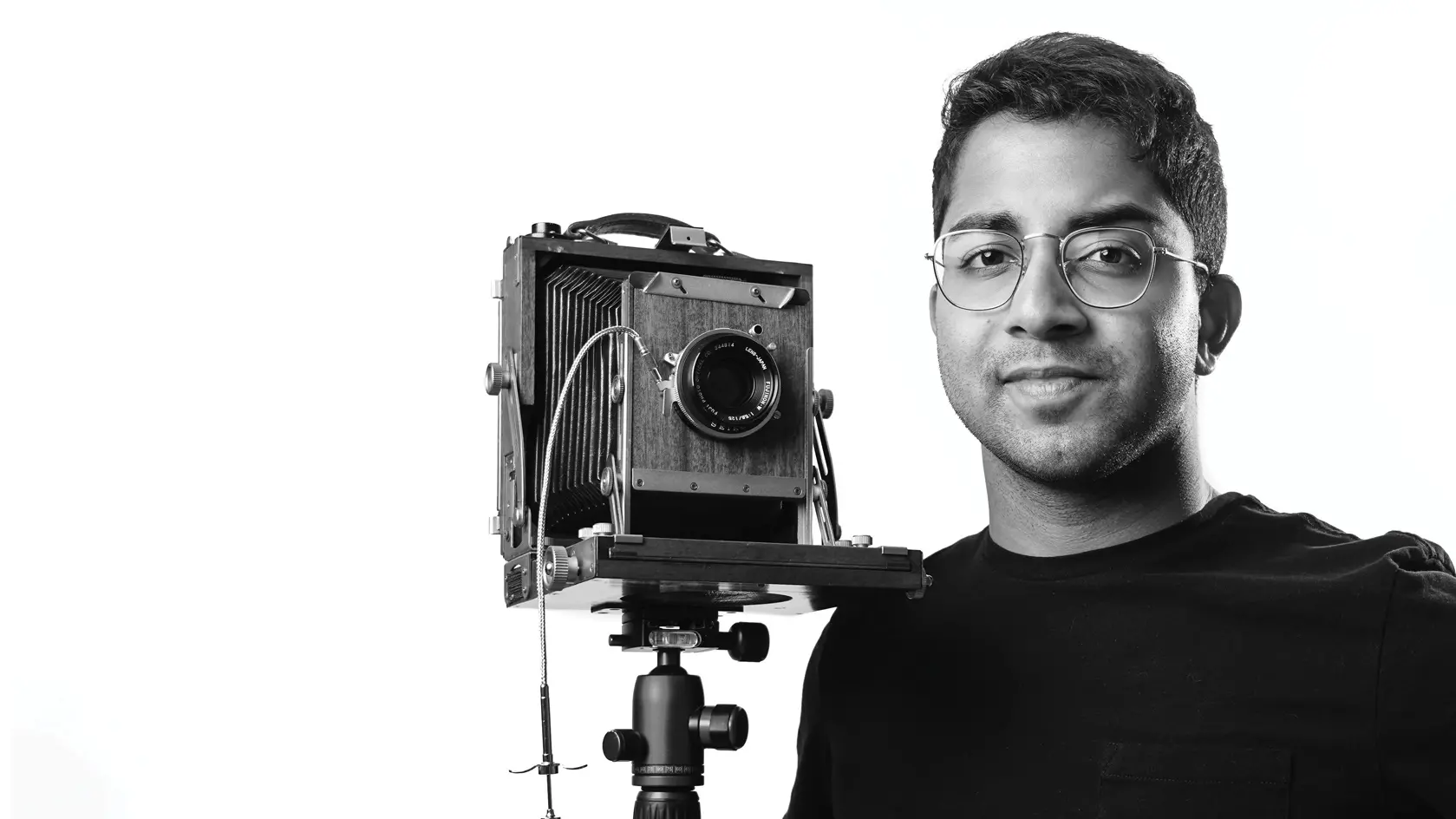 A young man of Indian descent stands beside a camera on a tripod and subtly smiles in a black and white self-portrait. He wears wire-rimmed glasses and his dark hair is short on the sides and a little curly on top.