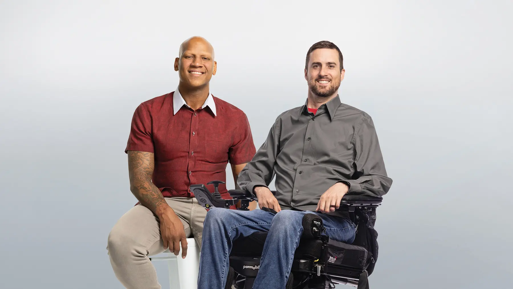 Two men sit next to each other smiling. On the left is a Black man on a stool wearing a short-sleeved button-down short that shows tattoos on his arms, a watch and shiny wedding ring. He casually rests his hands on his legs. On the right, a white bearded man in a motorized wheelchair wears a button-down shirt and a dark wedding ring.