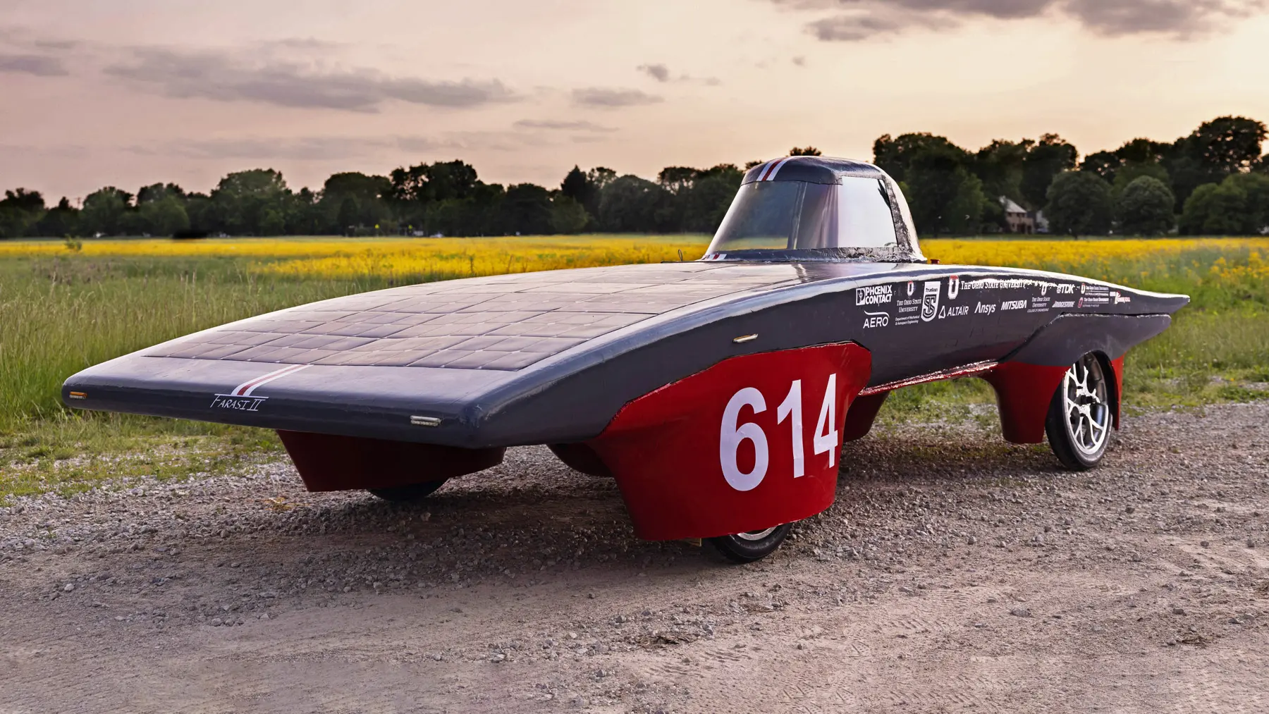 The Ohio State team’s solar car is scarlet and gray. There’s a one-person cockpit and the main body is softly arch-shaped, coated in solar panels and surprisingly shallow. A large 614 is emblazoned on the wheel cover closest to the camera. The car sits in front of a field during a sunset. 