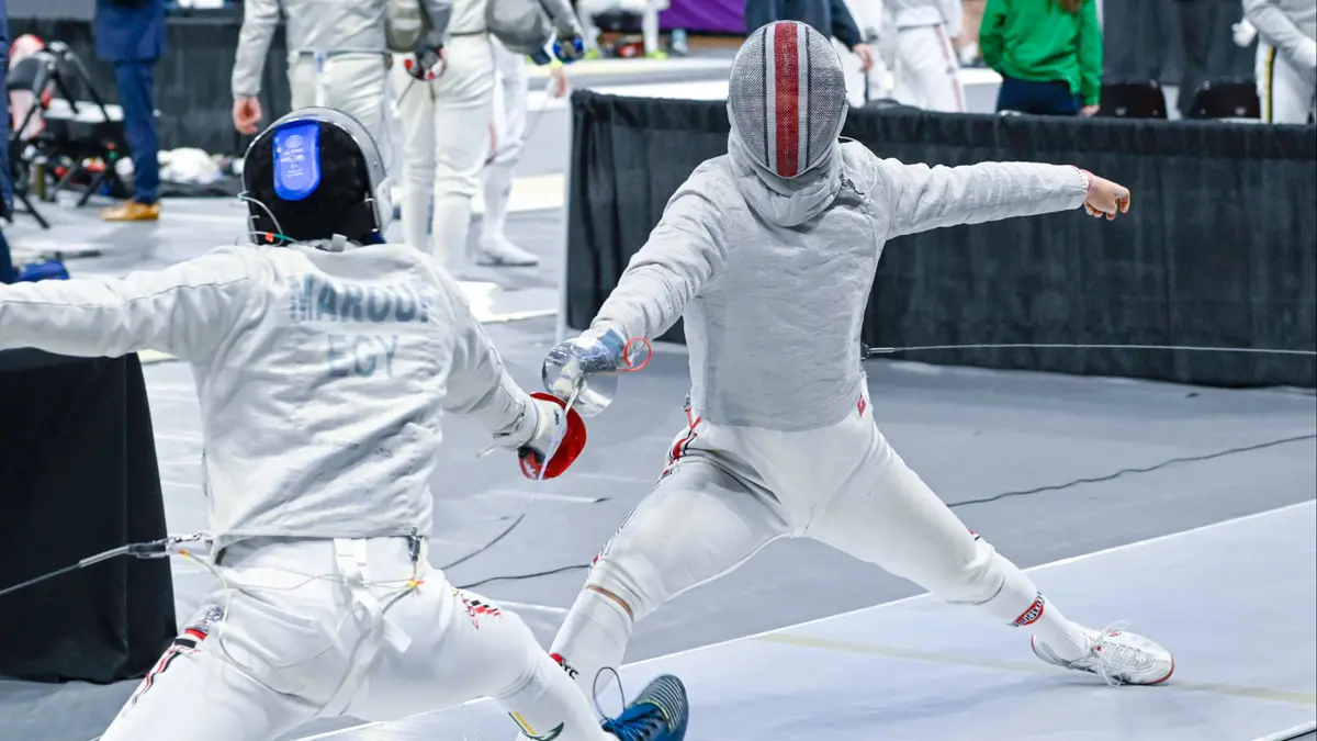 A fencer wearing a mask with Ohio State-colored stripes down the center lunges forward as he thrusts his sabre toward his opponent, who can only be seen from behind. Both wear padding and they’re at a competition. People behind them watch another match.