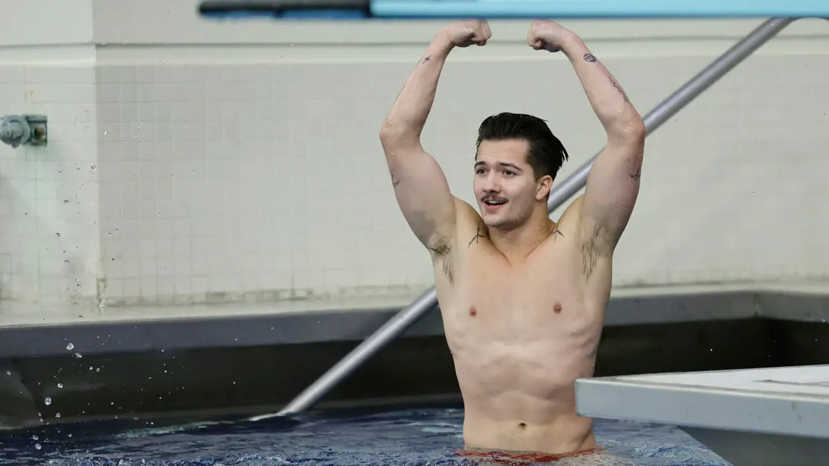 A shirtless young man with dark hair and a mustache raises his arms, as if halfway between a victory cheer and flexing his biceps. He’s looking at something off camera, perhaps a scoreboard or coach.
