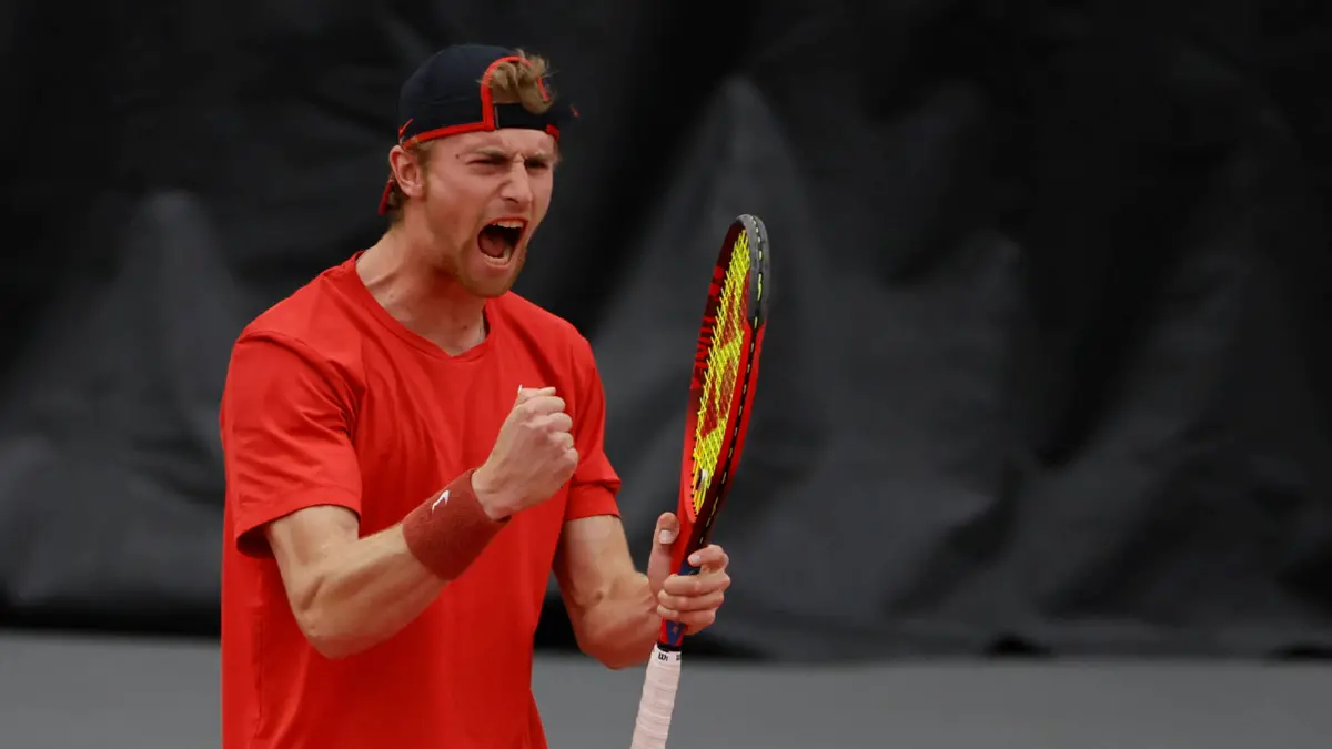 A young man in a backwards ballcap and T-shirt yells as he clenches a fist and holds his tennis racket. He looks intense, as if he just won a hard-fought volley.