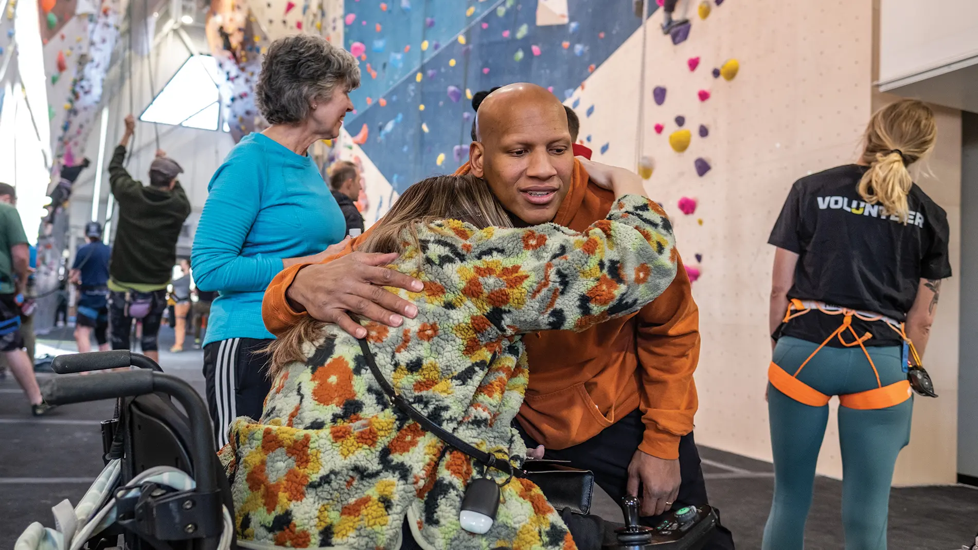 At a gym where a giant climbing wall is scattered with colorful hand holds, a Black man in a sweatshirt bends forward to hug a woman in a wheelchair. Her face is against his chest and can’t be seen, but he smiles as he pats her back. People behind them are focused on those climbing the wall. 