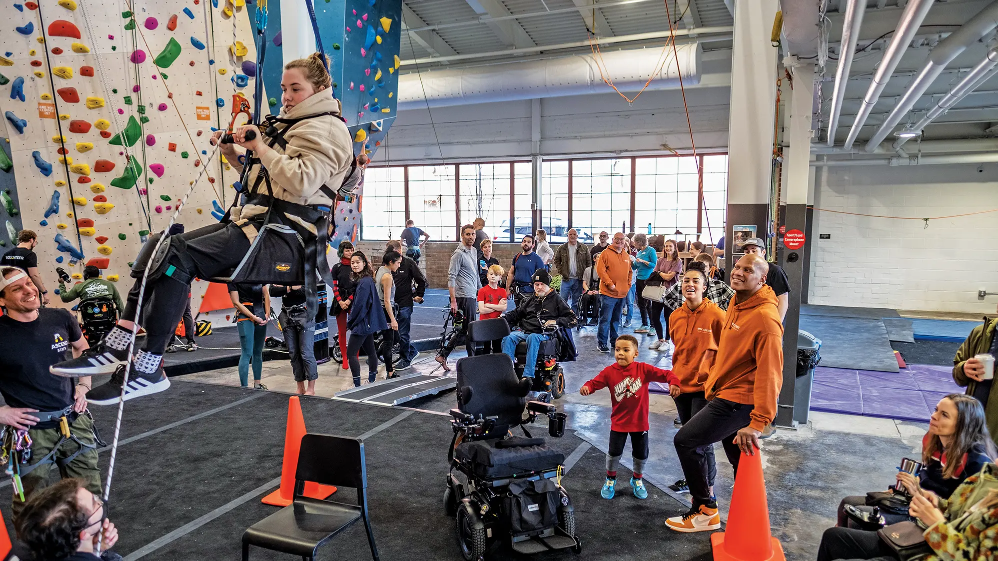 A white woman in a seat hoisted into the air holds a hand bar in front of her as she works to ascend a climbing wall. Lots of people fill the floor of the gym, including three watching her: a mom, dad and young child. The climber’s empty wheelchair sits in from of the family.